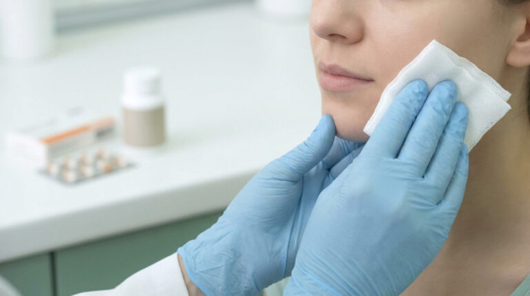 Hands in blue medical gloves gently press white gauze to a person's jaw. Blurred medical bottle and pills in background.