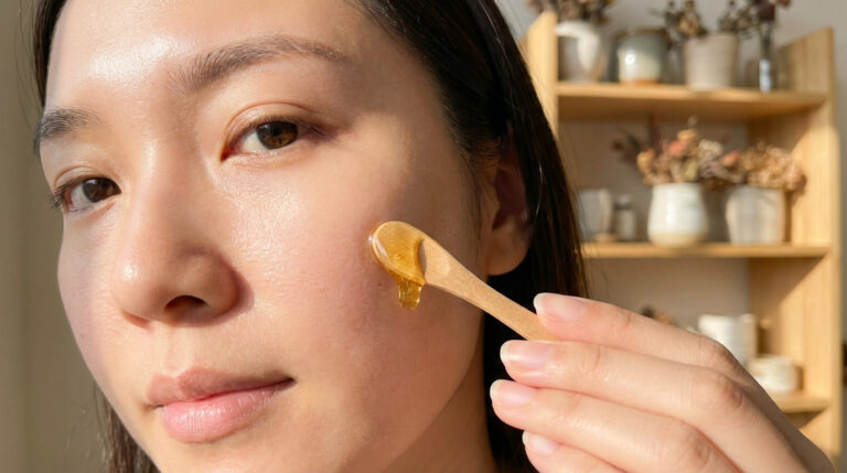 Close-up of a person's radiant face, a wooden applicator applies golden honey to smooth skin, blurred warm background.